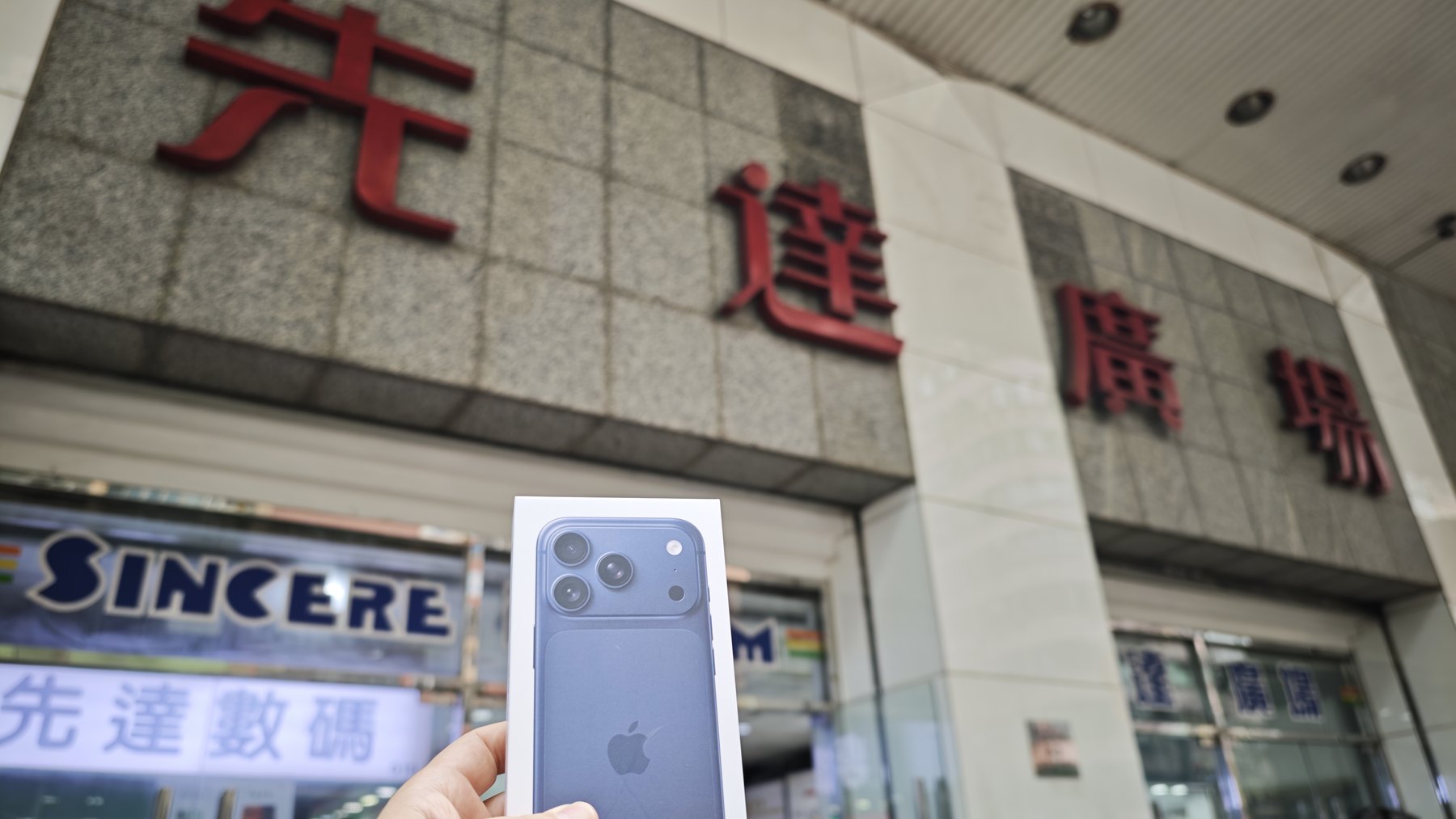 旺角・先達廣場の赤い看板の下で、買ったばかりの iPhone 18 の箱を持つ手