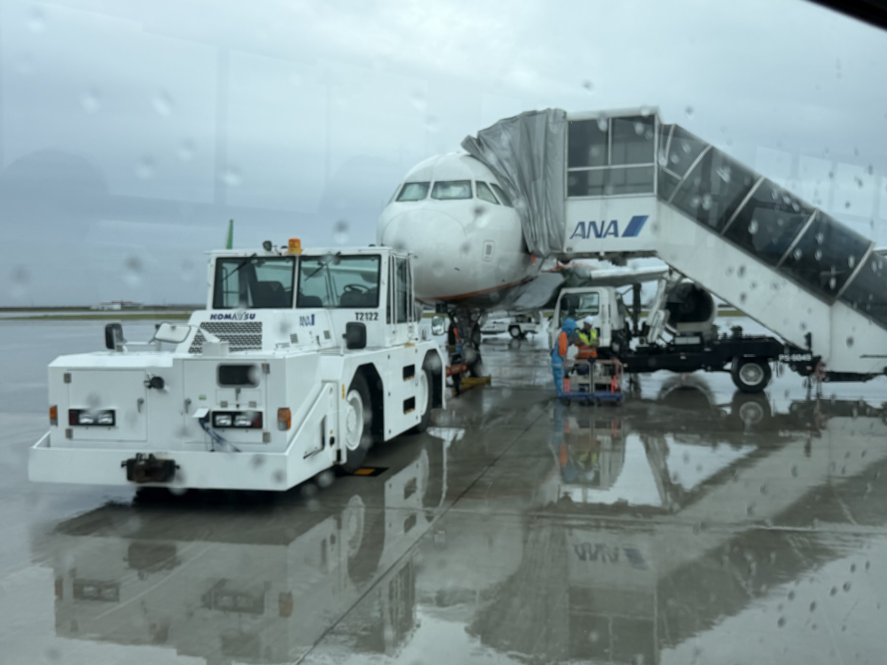 雨粒のついた窓越しの神戸空港の駐機場。ANAのボーディングブリッジにつながる機体と、白いトーイングトラクター