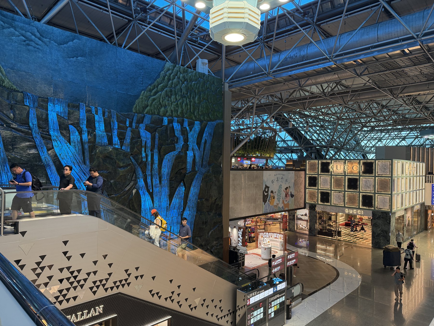 桃園空港の吹き抜け。青く光る滝を描いた岩壁のオブジェとエスカレーター、下の階に免税店が並ぶ
