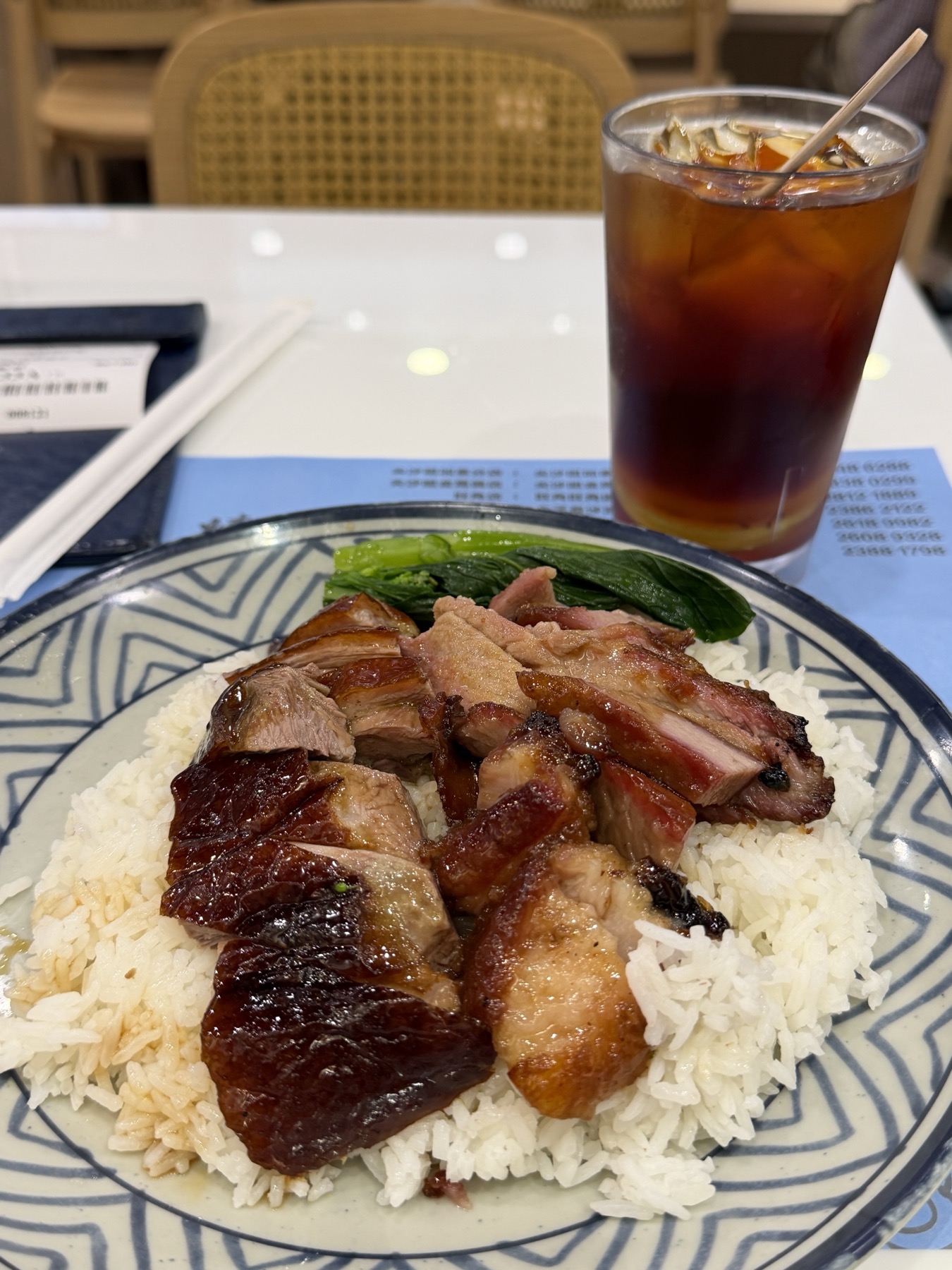 白飯の上に焼鵝と叉燒がハーフハーフで盛られた深皿、菜心が一本のせられ、奥に氷のたっぷり入った凍檸茶。プレースマットには支店の電話番号が並ぶ。