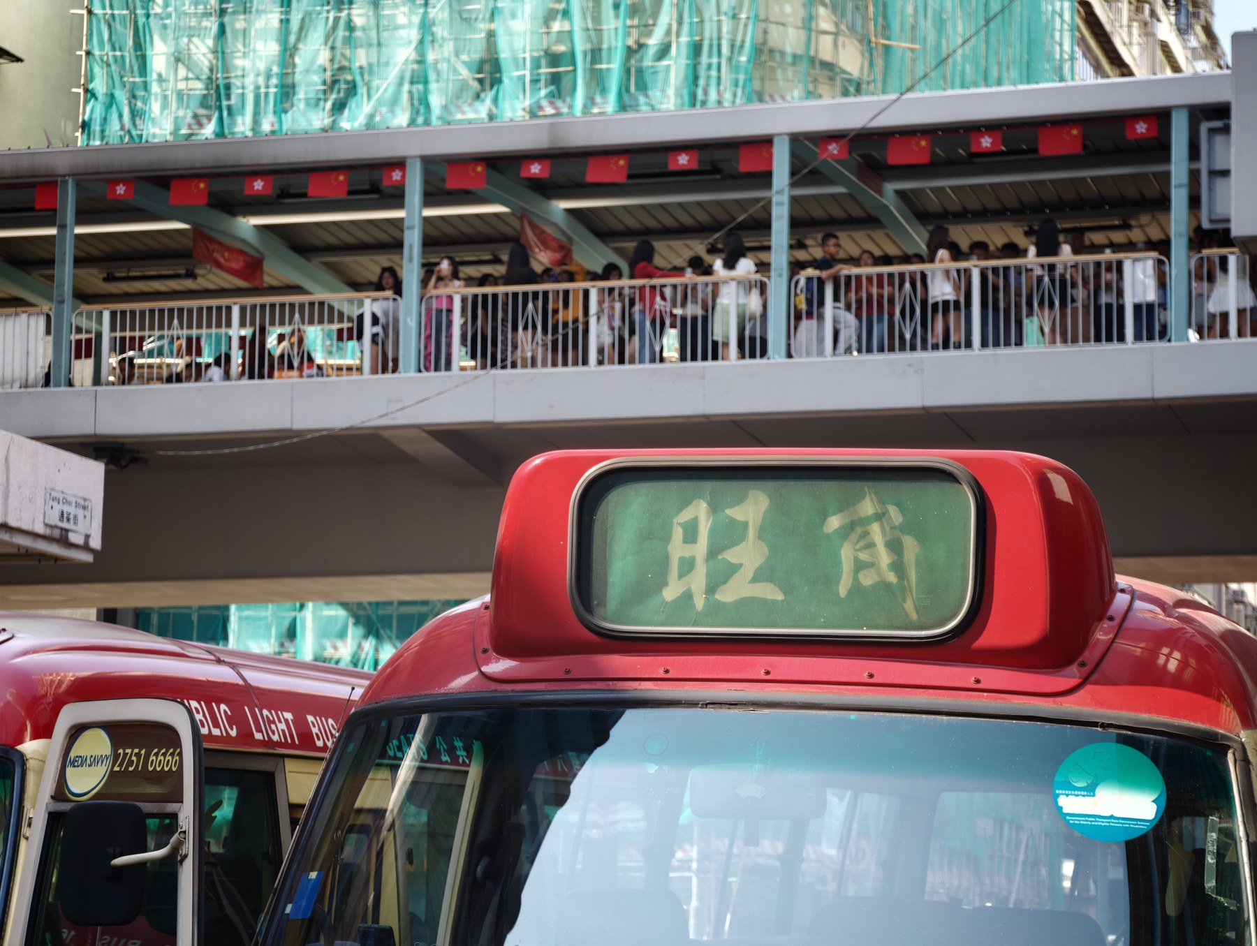 旺角行きの赤いミニバス越しに見上げた歩道橋。中国と香港の旗が並び、欄干沿いには写真を撮る大勢の人が集まっている