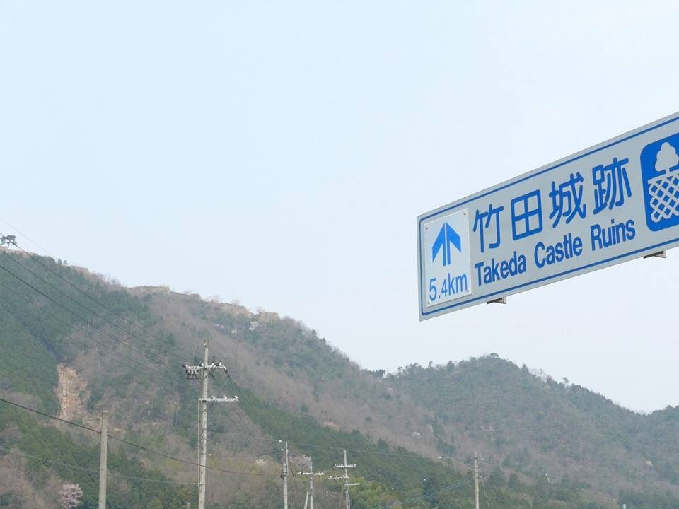 「竹田城跡 5.4km Takeda Castle Ruins」と書かれた青い道路標識と、その左手に見える山