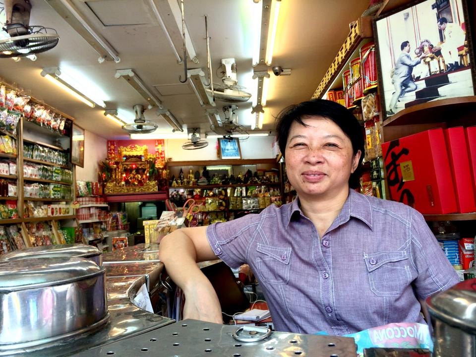 バンコク中華街の店のカウンターに立つ女性。棚には菓子や缶詰、奥に金色の祭壇が見える