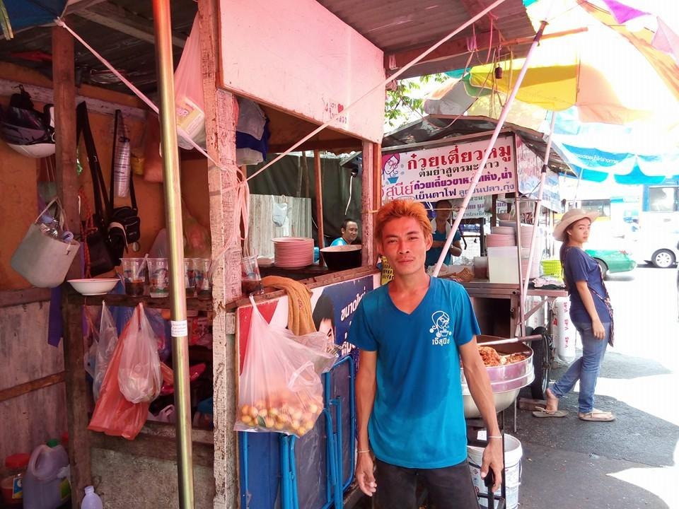 麺屋台の前に立つ青いシャツの店員。カメラに向かって穏やかに笑っている